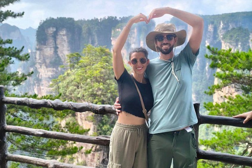 A couple making a heart shape with their hands, standing by a wooden railing with a mountainous landscape in the background. 4