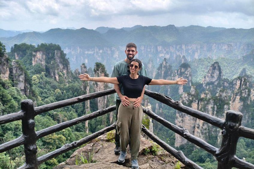 A couple standing on a wooden platform with outstretched arms, overlooking a mountainous landscape with steep cliffs and lush greenery. 1
