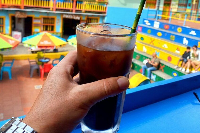 A hand holding a glass of iced tea with a straw, with a colorful outdoor seating area in the background. 2