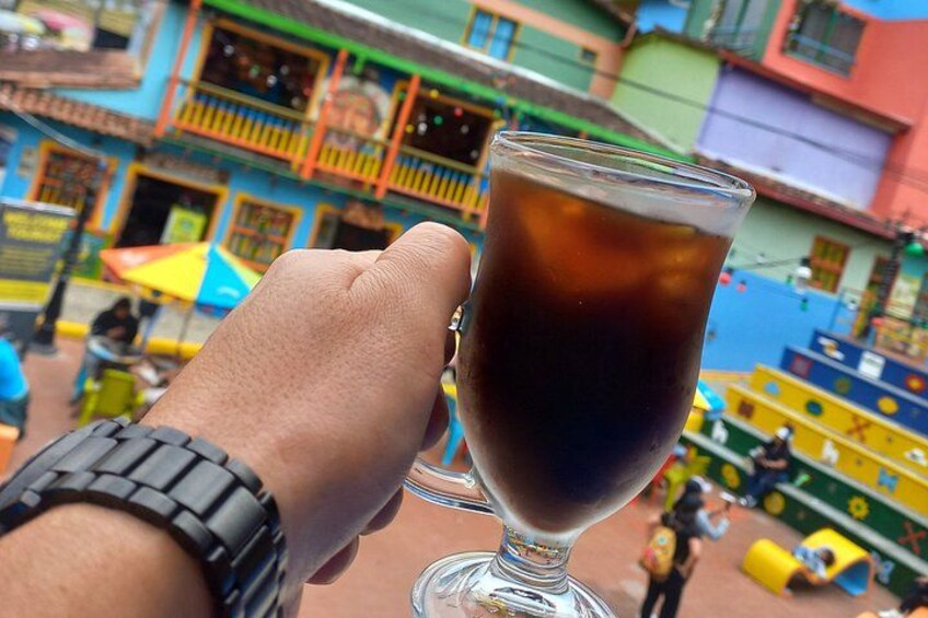 A hand holding a glass of dark liquid, possibly a beverage, with a colorful urban street scene in the background. 3