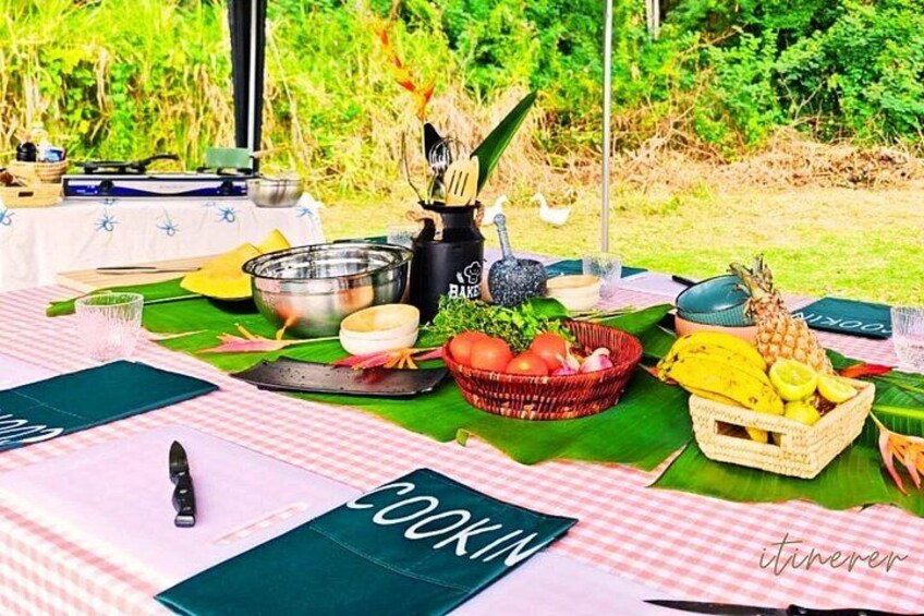 Un coin pique-nique avec une table recouverte d’une nappe à carreaux, un set de table vert sur lequel est écrit le mot « COOKIN’ », et divers aliments, notamment un panier rempli de fruits et de légumes. 1