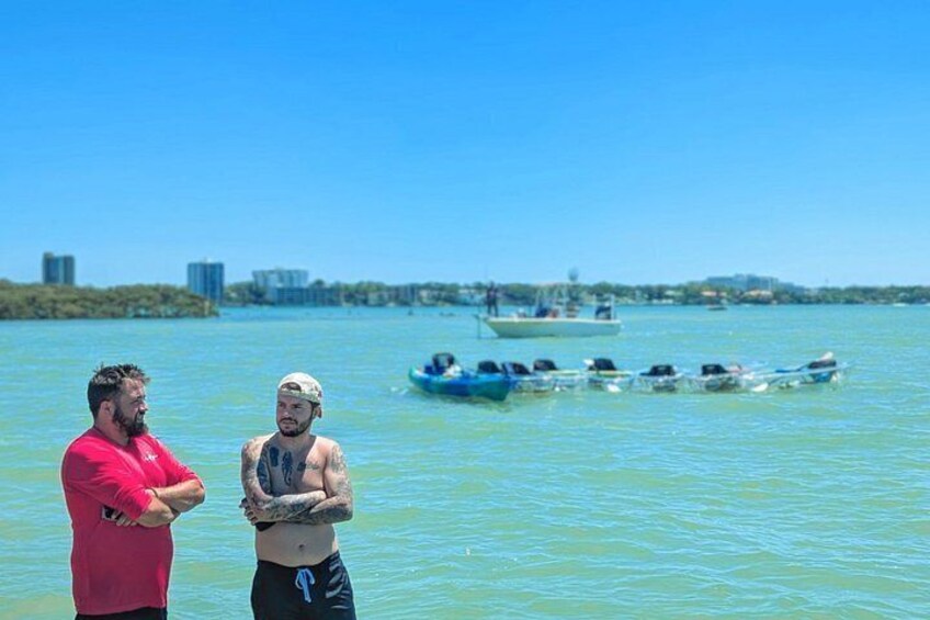 Deux personnes se tiennent près d’une étendue d’eau calme, avec une rangée de petits bateaux en arrière-plan. 1