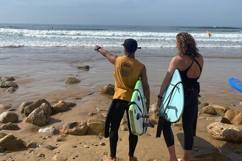 To personer på en stenet strand med surfbrædder, havnebølger og en svømmer i det fjerne. 5
