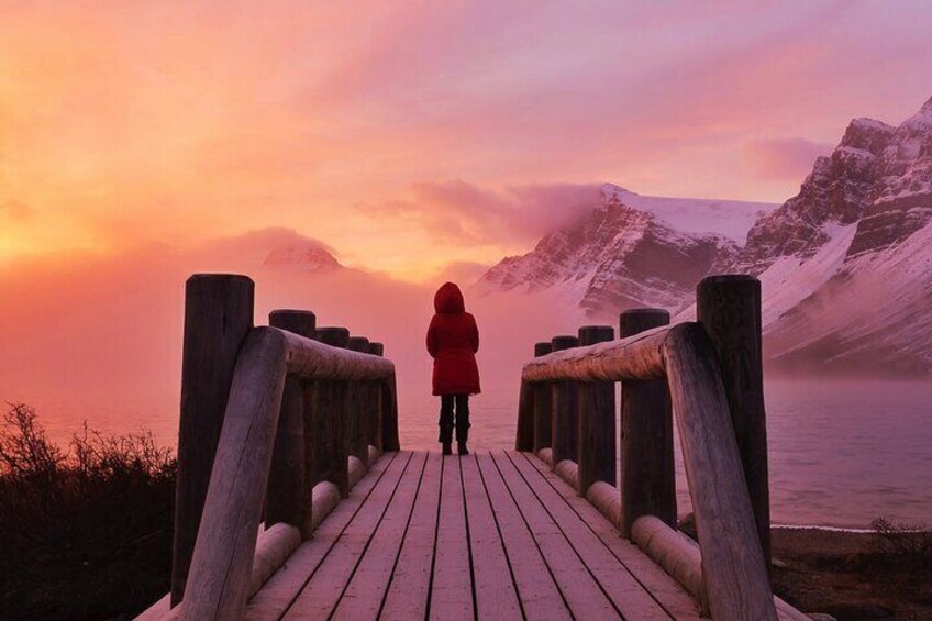 A person in a red jacket standing on a wooden boardwalk, overlooking a mountainous landscape during sunset. 1
