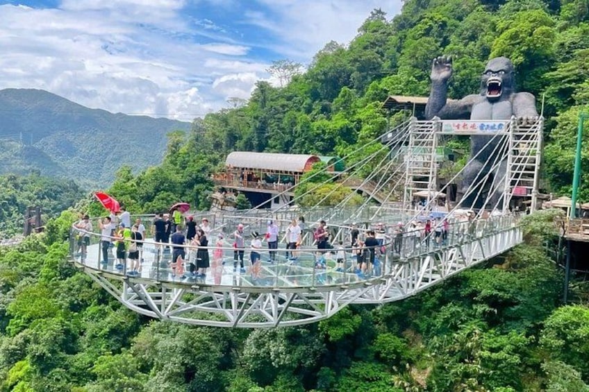 A glass bridge with a large gorilla statue, surrounded by lush greenery and mountains. 2