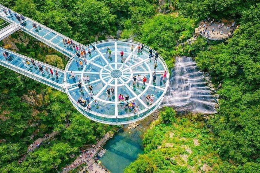 A glass bridge with people walking on it, surrounded by lush greenery and a waterfall. 1