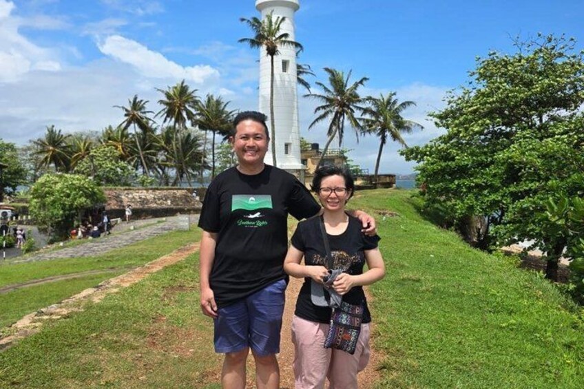 Two people standing in front of a lighthouse with palm trees and a grassy path in the background. 2