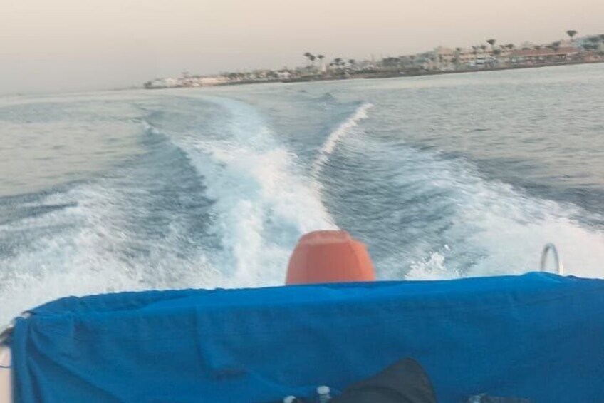 Un bote navegando por el agua, dejando una estela, con un toldo azul en la cubierta y una costa con edificios al fondo. 3