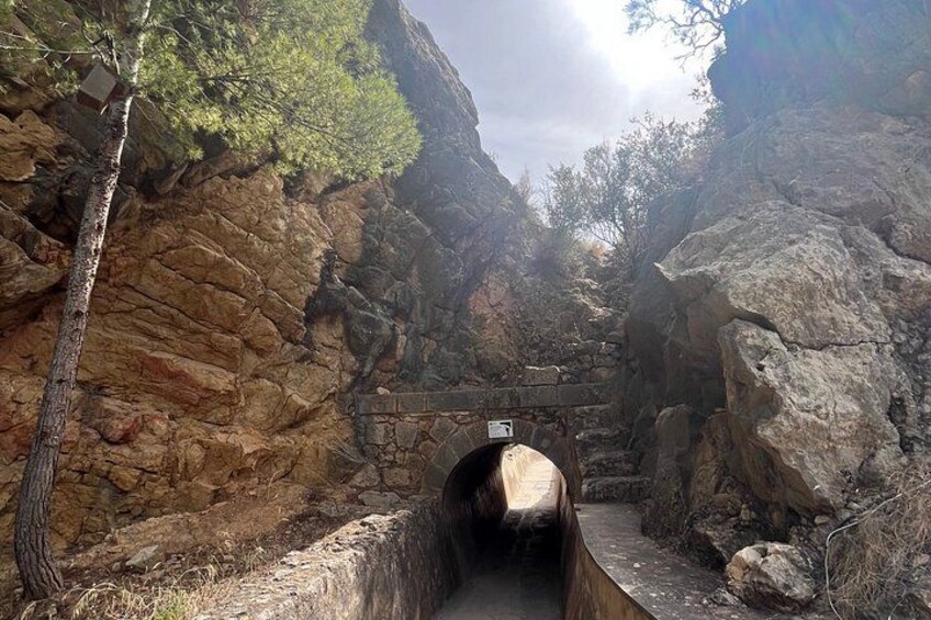 A narrow stone tunnel with a wooden walkway, surrounded by rocky cliffs and sparse vegetation. 4