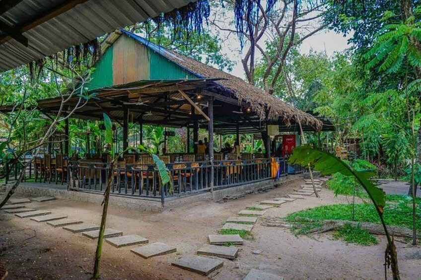 An outdoor restaurant with a thatched roof, surrounded by lush greenery and a dirt path. 2