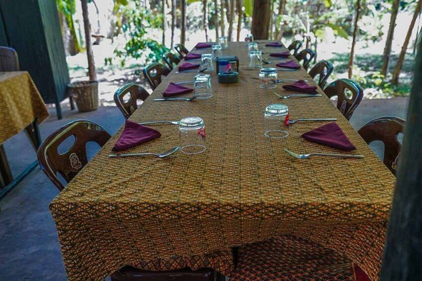 A long table set for dining outdoors with chairs, a woven tablecloth, and purple napkins. 5