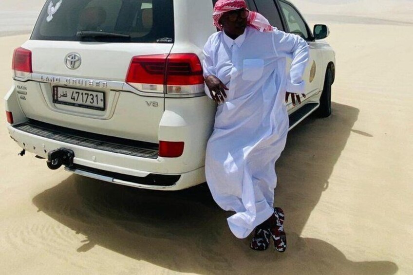 Un hombre con vestimenta tradicional apoyado en un Toyota Land Cruiser blanco en un desierto. 1