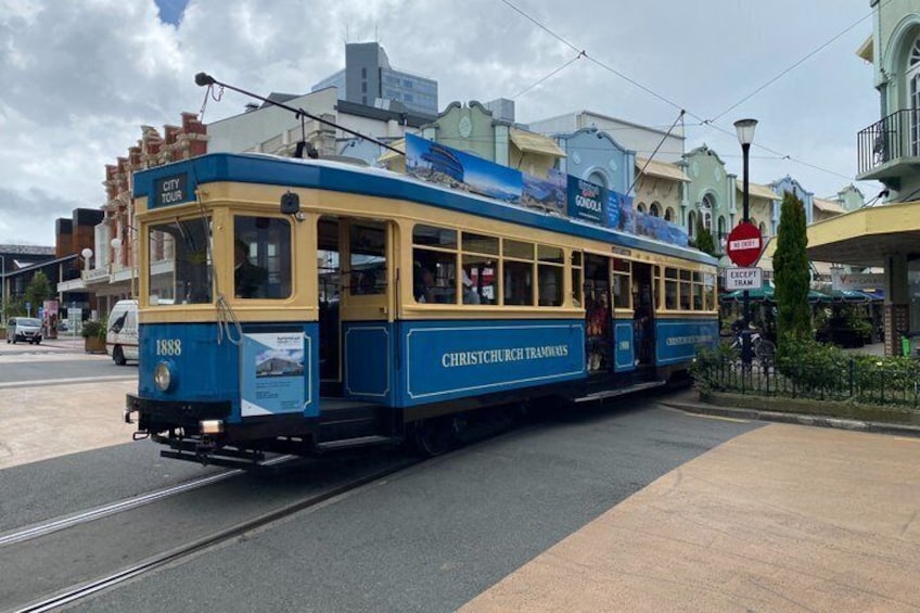 En blå og gul sporvogn med teksten 'City Tour' og 'Christchurch Tramways' parkeret på en gade med bygninger i baggrunden. 1
