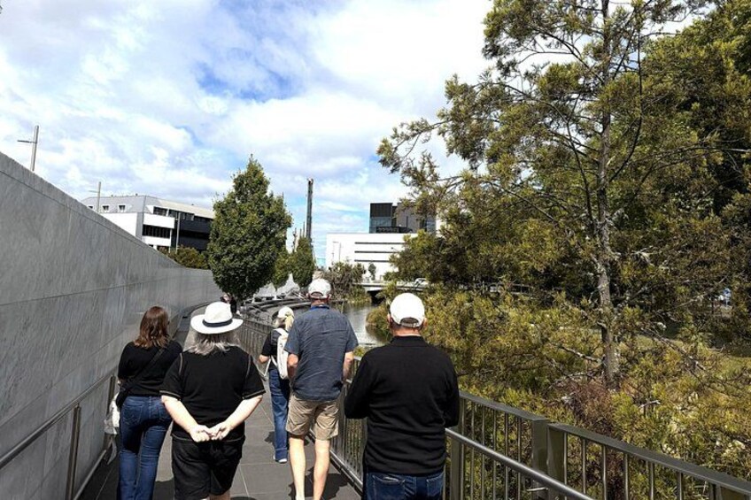A group of people walking on a concrete path beside a river, with trees and buildings in the background. 3