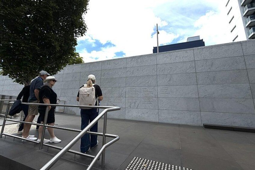 A group of people standing near a wall with inscriptions, a metal railing, and a tree in the background. 2