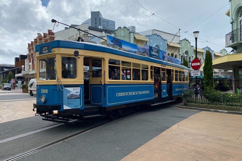 A blue and yellow tram with 'City Tour' and 'Christchurch Tramways' written on it, parked on a street with buildings and a sign that says 'No Entry' in the background. 2
