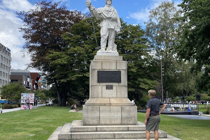 A statue in a park with a plaque, grass, trees, and buildings in the background. 5
