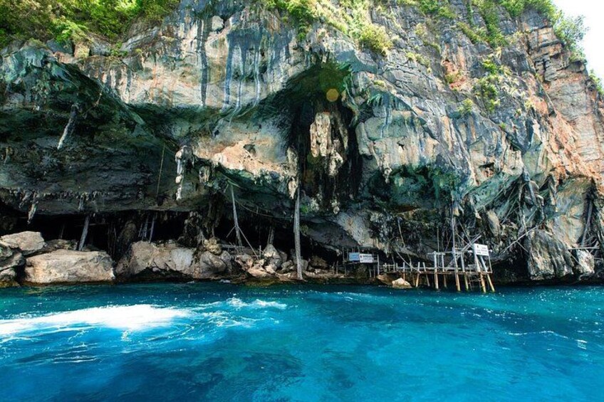 Un acantilado rocoso con la entrada de una cueva, agua azul cristalina y pasarelas de madera. 4