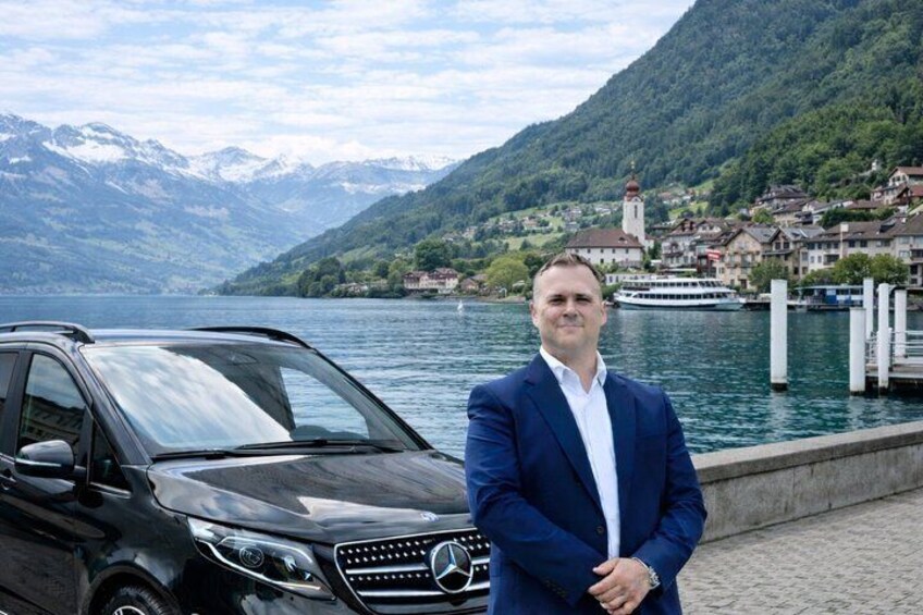 Een man in een blauw pak staat naast een zwarte Mercedes-Benz SUV bij een meer, met bergen op de achtergrond. 1