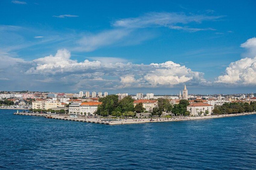 A coastal cityscape with a prominent church tower, waterfront promenade, and dense urban buildings. 1