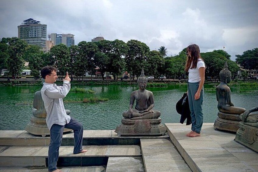 A person taking a photo of a statue, another person standing next to a statue, and a body of water with trees in the background. 4