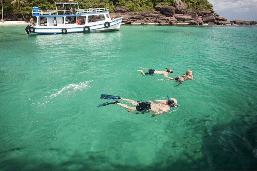 En gruppe mennesker snorkler i klart turkis vand, en båd i baggrunden og en klippefyldt kystlinje med vegetation. 2