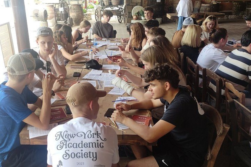 Un gruppo di persone sedute a un tavolo di legno all'aperto, impegnate a scrivere o leggere. 2