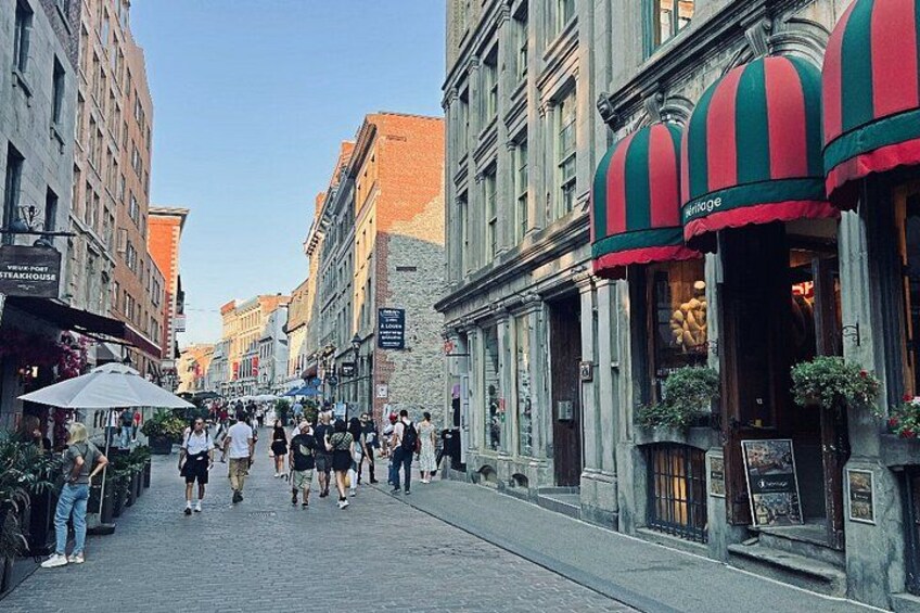 Eine Kopfsteinpflasterstraße gesäumt von historischen Gebäuden, ein Restaurant mit Streifenmarkisen und Fußgänger, die entlanggehen. 1
