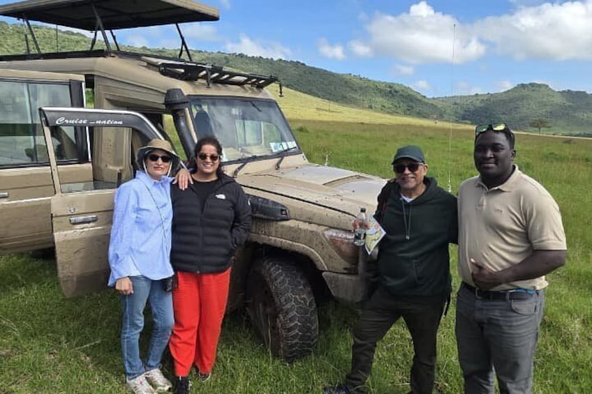 Un grupo de personas de pie junto a un vehículo safari en un campo cubierto de hierba, con colinas al fondo. 1