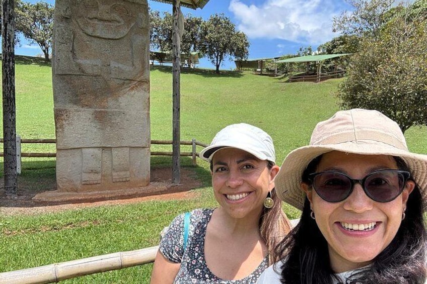 Two people standing in front of a stone monument with a grassy field and trees in the background. 4