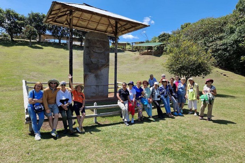 A group of people standing in a grassy area with a stone monument and a wooden shelter. 3