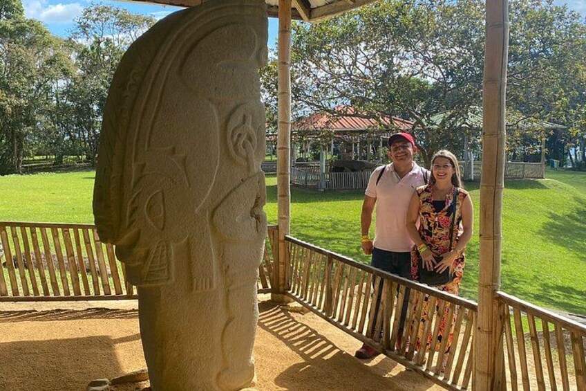 A stone monument with carvings, a wooden fence, and a grassy area with trees in the background. 2