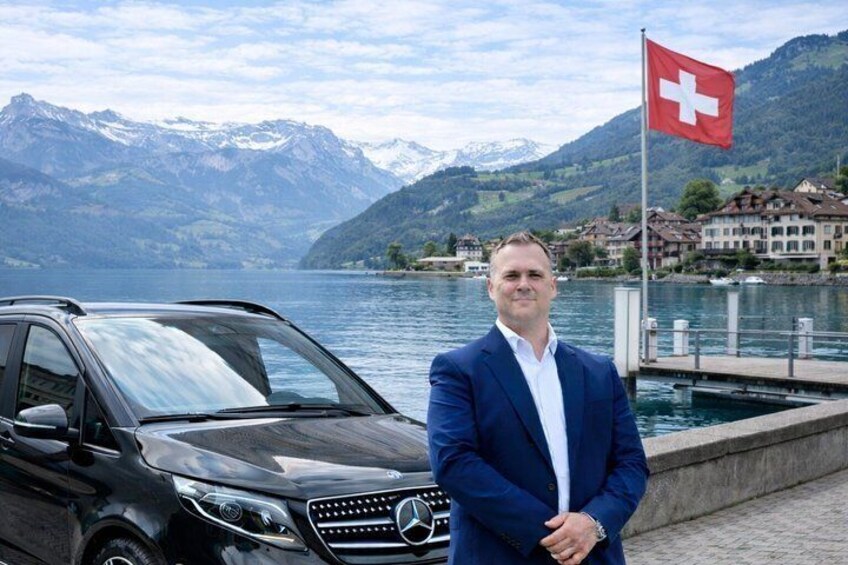 Un hombre con traje azul parado junto a un SUV Mercedes-Benz negro frente a un lago, con montañas al fondo y una bandera suiza. 1