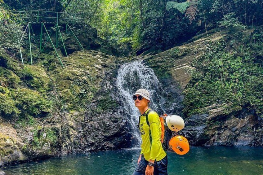 A person in a yellow shirt and orange vest standing by a waterfall in a forested area. 3