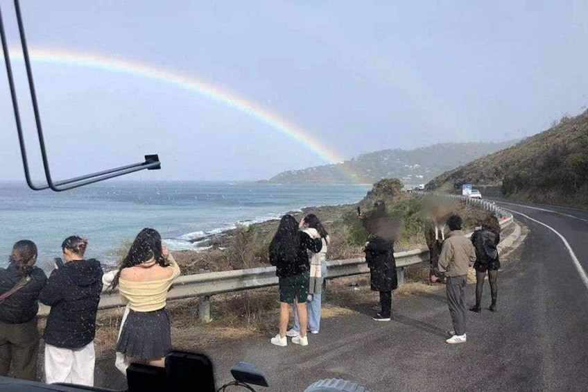 A group of people observing a rainbow over the ocean from a roadside. 3