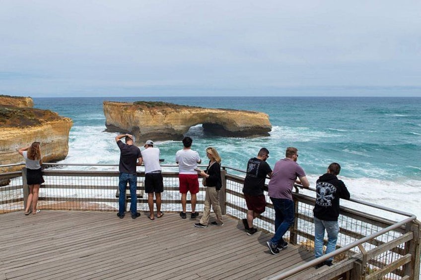 A wooden boardwalk with people observing the ocean and a large rock formation. 4