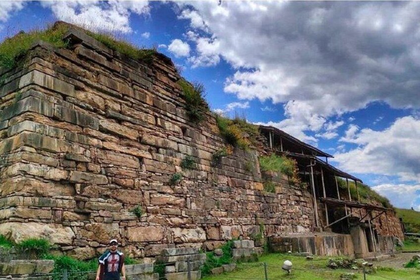 A stone wall with a wooden structure on top, a person standing in front, and a clear sky with clouds. 4
