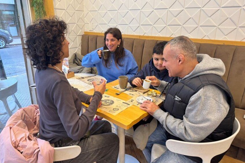En grupp människor sitter vid ett bord på ett kafé, med en vägg med geometriskt mönster i bakgrunden. 3