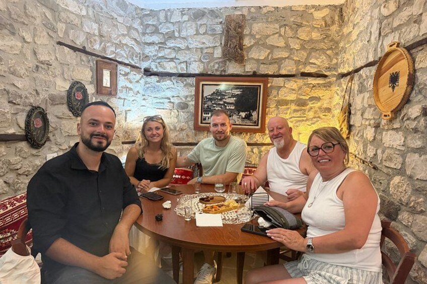 Un gruppo di persone sta cenando a un tavolo rotondo in una sala con pareti in pietra e un quadro appeso al muro. 2