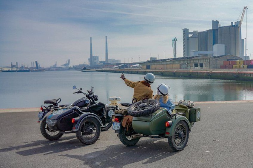 Två motorcyklister på en asfalterad väg, med en vattenyta och industriella byggnader i bakgrunden. 3