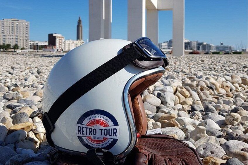 En hvid hjelm med brun rem og mærket 'RETRO TOUR', placeret på en stenet overflade med bygninger i baggrunden. 5