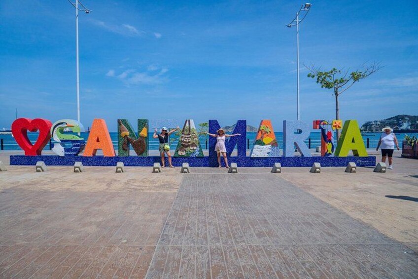 A colorful sign with the word 'CANAMARINA' and a heart symbol, with a clear blue sky and a body of water in the background. 2