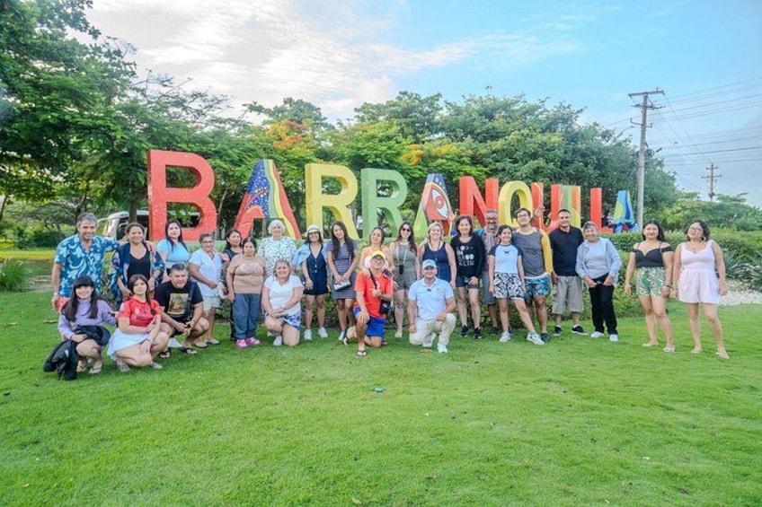 A group of people posing in front of large colorful letters spelling 'BARRANQUIL' on a grassy field with trees in the background. 1