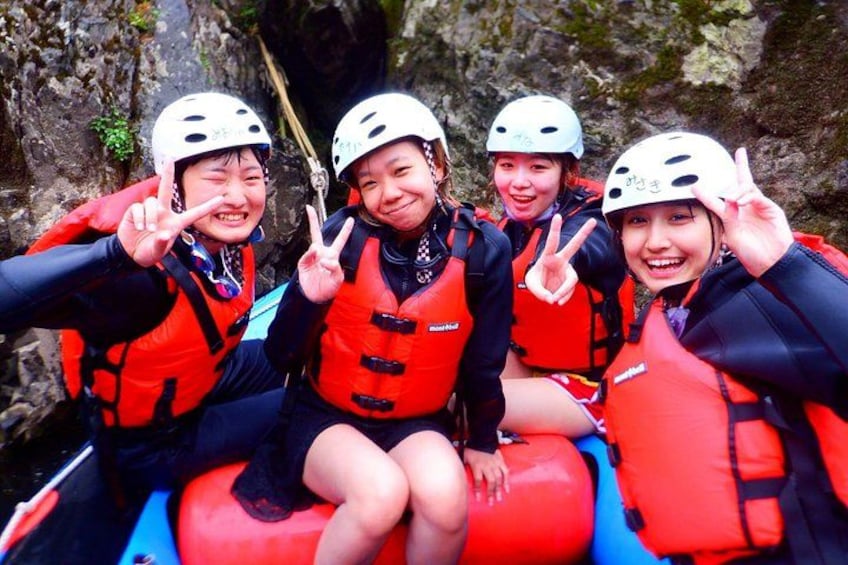 Four people in a raft wearing life jackets and helmets, with a rocky terrain in the background. 4