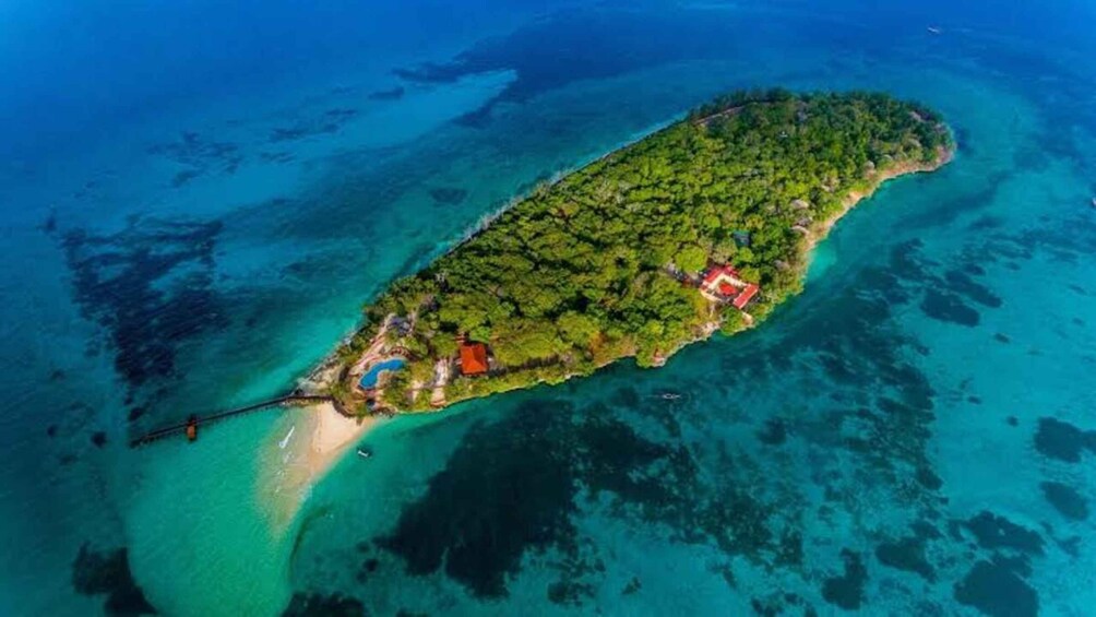 An aerial view of a tropical island with clear turquoise waters, dense green vegetation, and a few buildings. 1