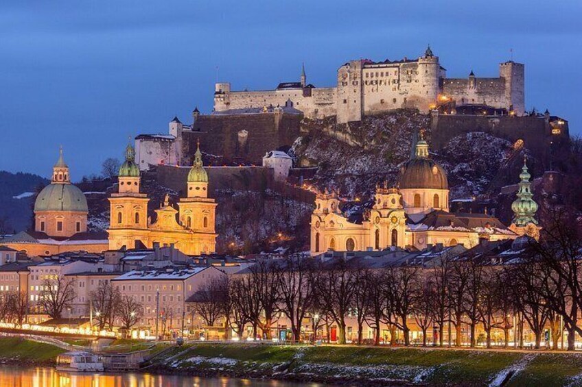 A historic cityscape with a castle on a hill, illuminated buildings, and a river in the foreground. 3