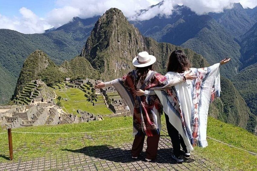 Two people standing on a paved area with a view of Machu Picchu and surrounding mountains. 2