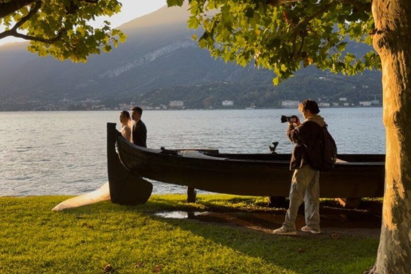 Ein Paar sitzt an einem See auf einer Bank, im Hintergrund ein Boot und eine Person macht ein Foto. 3