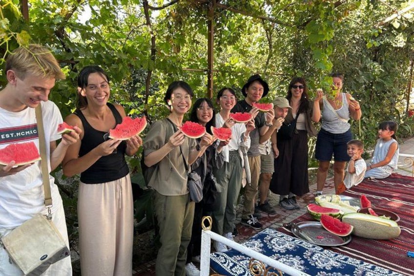 Un groupe de personnes, à l’extérieur, tenant des tranches de pastèque, devant une table sur laquelle sont disposées des demi-pastèques et un tissu bleu à motifs. 3