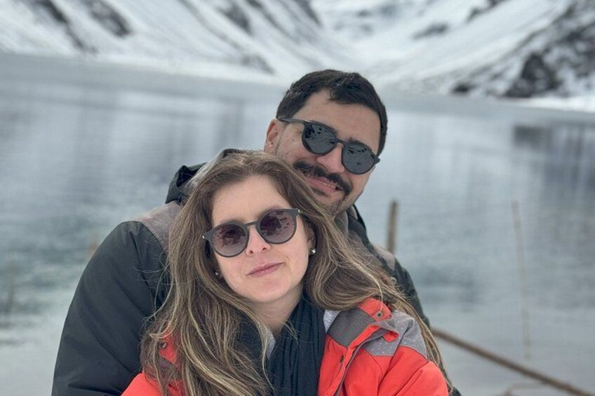 Eine Person mit roter Jacke und Sonnenbrille steht vor einem zugefrorenen See, im Hintergrund sind schneebedeckte Berge zu sehen. 2